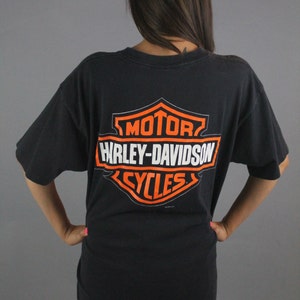 Puede incluir: Camiseta negra con un gran logotipo de Harley-Davidson naranja y blanco en la espalda. El logotipo incluye las palabras "Motor", "Harley-Davidson" y "Cycles".