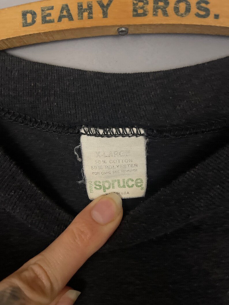 Puede incluir: Una etiqueta negra con costuras blancas, que dice "X-Large", "50% Algod&oacute;n", "50% Poli&eacute;ster", "Para el cuidado, consulte el reverso", y "spruce" en letras verdes. La etiqueta est&aacute; hecha en los Estados Unidos.