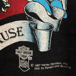 May include: Black t-shirt with a Harley Davidson logo and a graphic of a stylized American flag with a banner that reads "USE". The text "&copy; 1987 Harley Davidson, Inc. Mfd. by Speed Limit Seventy" is printed on the bottom of the shirt.