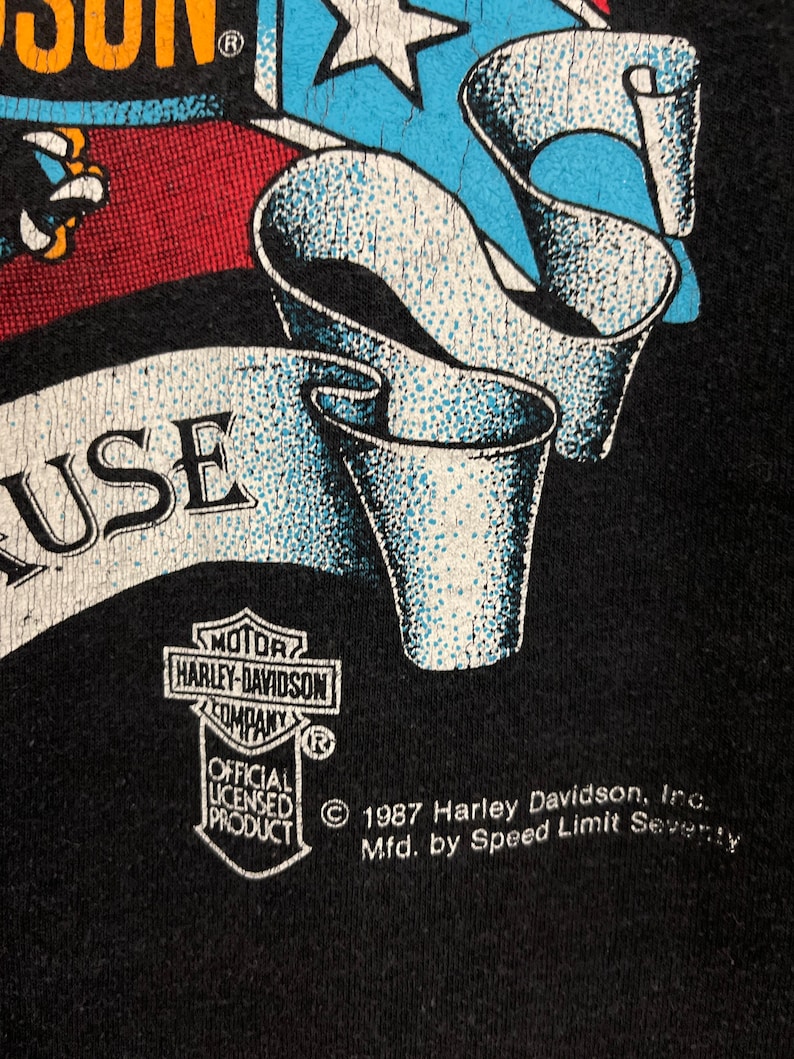 May include: Black t-shirt with a Harley Davidson logo and a graphic of a stylized American flag with a banner that reads "USE". The text "&copy; 1987 Harley Davidson, Inc. Mfd. by Speed Limit Seventy" is printed on the bottom of the shirt.