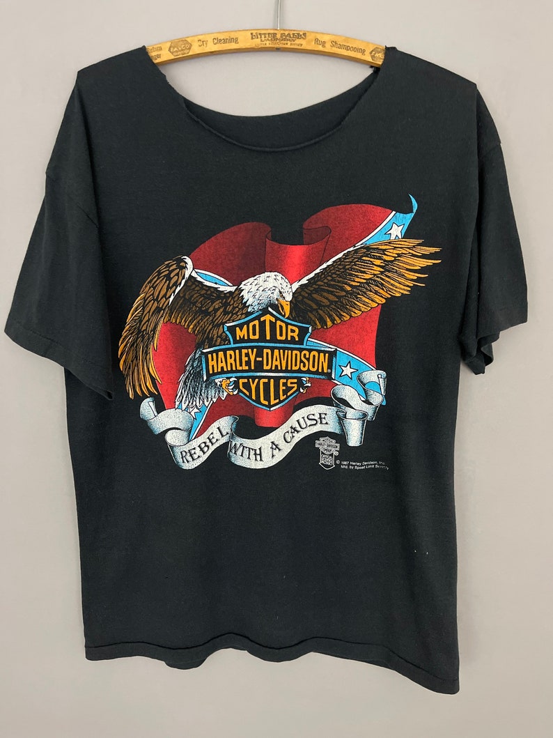 May include: Black t-shirt with a graphic of an eagle holding a Harley-Davidson logo with the text "Rebel with a Cause" and "Motor Harley-Davidson Cycles".