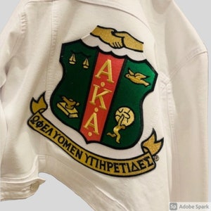 May include: White denim jacket with an embroidered green, gold, and red crest featuring the letters "AKA" and the Greek phrase "ΦΕΛΟΥΜΕΝ ΥΤΗΡΠΕΤΙΔΕΣ".