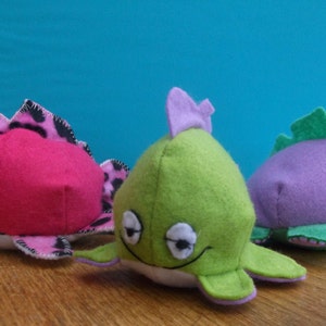 May include: Three plush toys shaped like fish. The first is pink with black and white spots, the second is green with white eyes, and the third is purple with green fins and white eyes.