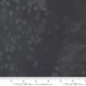 May include: Dark gray fabric with a subtle white speckled pattern. The fabric is textured and has a slight sheen. The fabric is measured in inches with the text "moda" printed on the ruler.