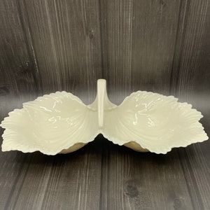 May include: A white porcelain serving dish with two sections and a handle. The dish is shaped like two leaves and has a raised design.