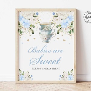 Op de afbeelding: Een blauw en wit printbaar bord met een bloemenontwerp en gestapelde theekopjes. Het bord zegt "Baby's zijn zoet" en "Neem gerust een traktatie".