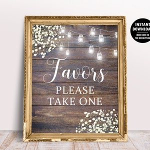 May include: A rustic wooden sign with a gold frame and white text that reads "Favors Please Take One". The sign is decorated with white flowers and string lights.