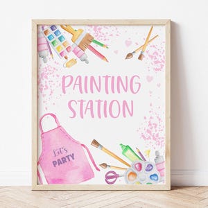 May include: A printable sign for a painting station at a party. The sign is pink with white polka dots and features a pink apron with the words "Let's Party" on it. The sign also includes watercolor paints, paintbrushes, and other art supplies.