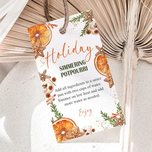 May include: A rectangular tag with the words "Holiday Simmering Potpourri" in orange and green script. The tag features illustrations of orange slices, gingerbread cookies, and greenery. Instructions for use are printed below.