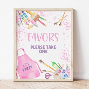 Puede incluir: Un cartel rosa y blanco con las palabras "Favors Please Take One" y un delantal rosa con las palabras "Let's Party". El cartel está decorado con pinceles de acuarela, pintura y otros materiales de arte.