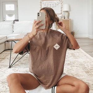 May include: A woman wearing a brown oversized t-shirt with a white graphic design on the front. She is sitting on a rug with her legs crossed and wearing white socks with black stripes.