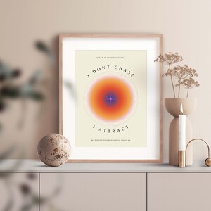May include: A framed wall art print with a cream background featuring a central orange and purple gradient circle with a star icon. The text reads: SPEAK IT INTO EXISTENCE, I DONT CHASE, I ATTRACT, and MANIFEST YOUR DEEPEST DESIRES in black lettering.