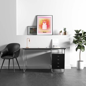 May include: A framed wall art print featuring a stylized pink and yellow gradient background with a central white vase shape and a vibrant orange flower, with the word bloom printed in black text at the bottom of the rectangular composition.