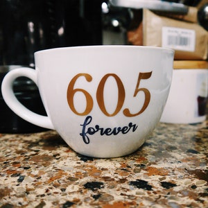 May include: White ceramic coffee mug with gold lettering that reads "605 forever".