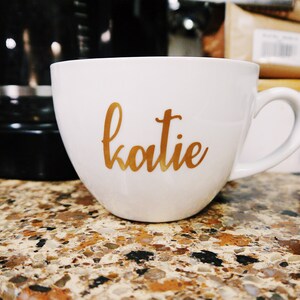 May include: White ceramic coffee mug with a gold inscription that reads "katie".