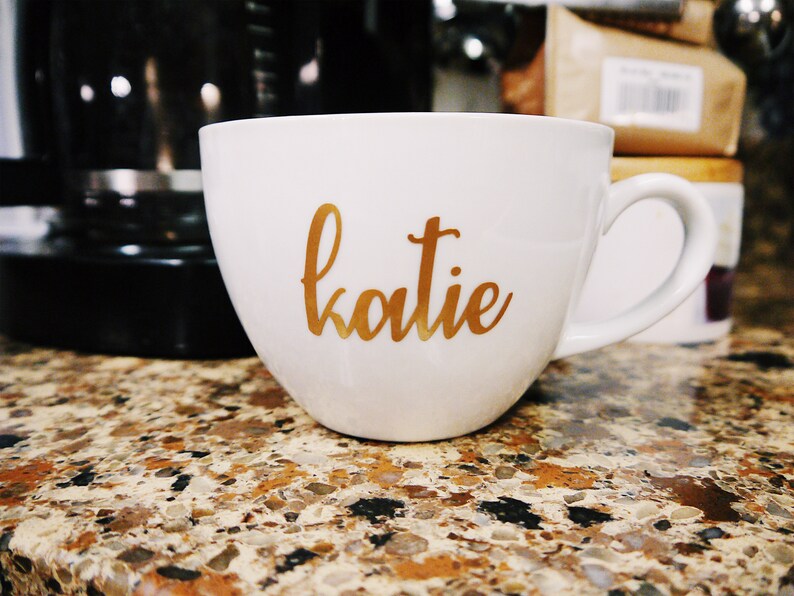 May include: White ceramic coffee mug with a gold inscription that reads "katie".