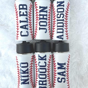 May include: White water bottles with black lids and the names CALEB, JOHN, ADDISON, NIKO, BROCK, and SAM in navy blue. Each bottle has red baseball stitching design on the sides.
