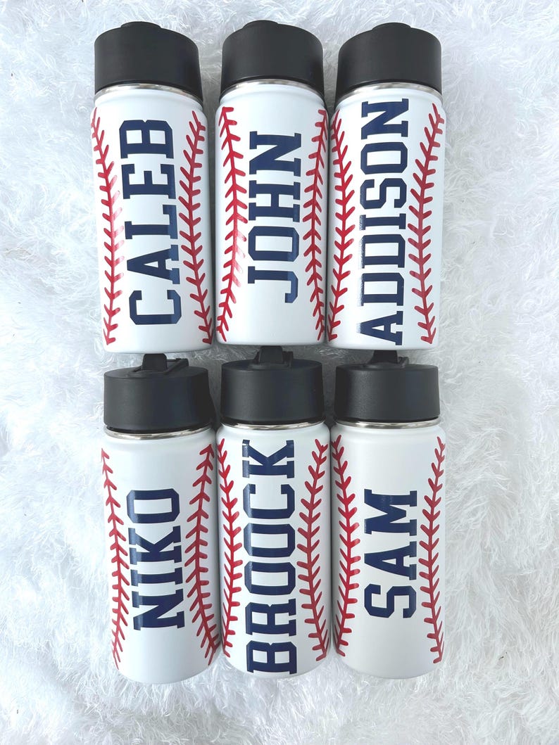 May include: White water bottles with black lids and the names CALEB, JOHN, ADDISON, NIKO, BROCK, and SAM in navy blue. Each bottle has red baseball stitching design on the sides.