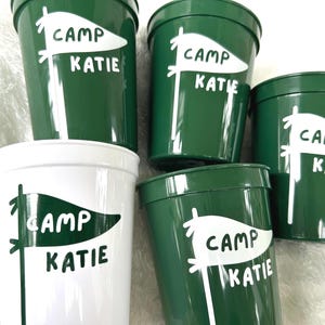 May include: Five green and white plastic cups with the text "Camp Katie" and a white flag design.