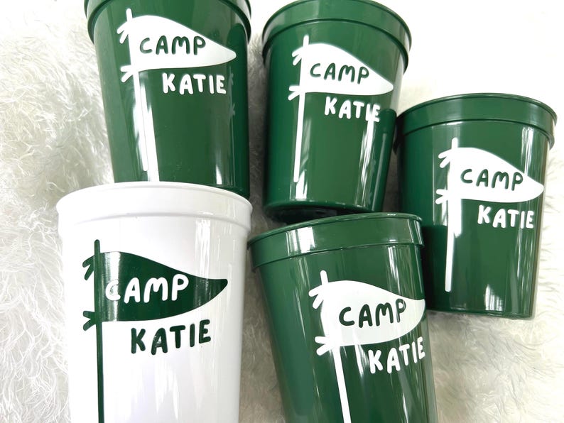 May include: Five green and white plastic cups with the text "Camp Katie" and a white flag design.