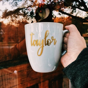 Peut inclure: Une tasse en c&eacute;ramique grise avec une inscription dor&eacute;e "Taylor". La tasse est tenue par une main.