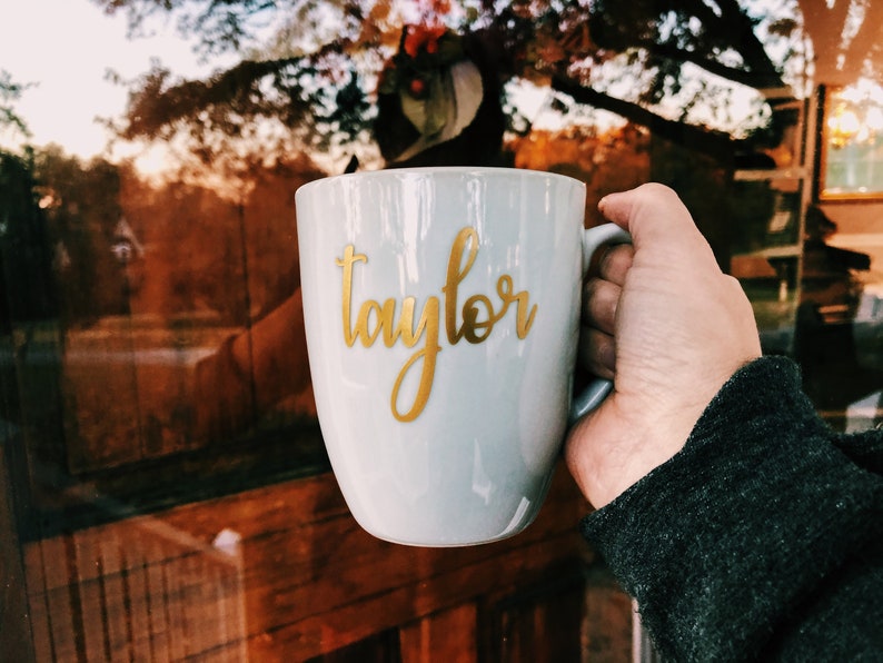 Peut inclure: Une tasse en c&eacute;ramique grise avec une inscription dor&eacute;e "Taylor". La tasse est tenue par une main.