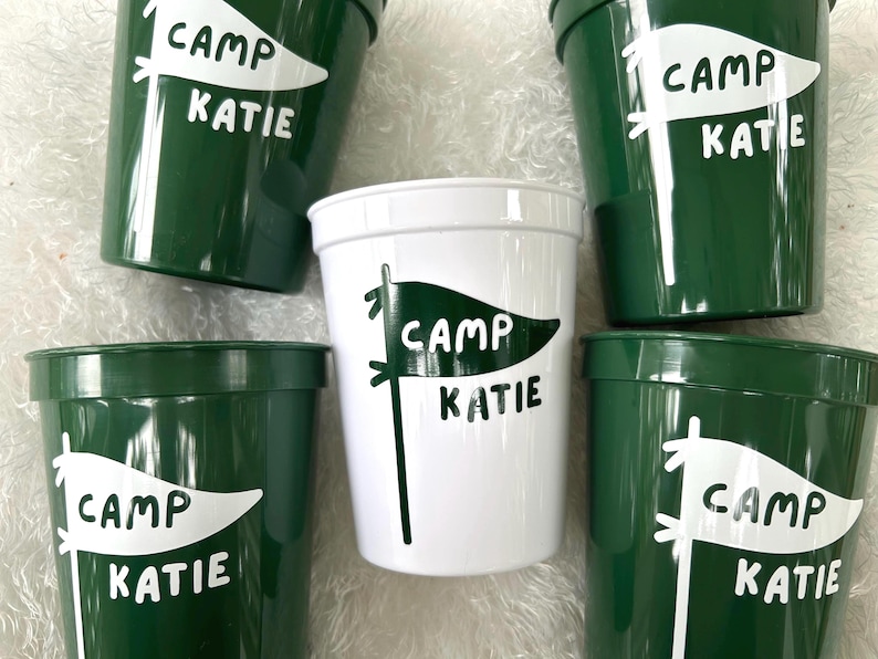 May include: Five green plastic cups with white lettering. Each cup has a white flag with the text "Camp Katie" on it.
