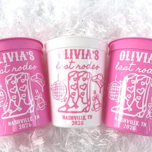 May include: Three plastic cups, two pink and one white, with a rodeo theme. The cups feature the text "OLIVIA'S last rodeo" and a graphic of cowboy boots, a disco ball, and a cowboy hat. The text "NASHVILLE, TN 2026" is also included.