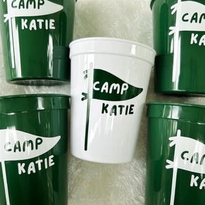 May include: Five green plastic cups with white lettering. Each cup has a white flag design with the text "Camp Katie" on it.