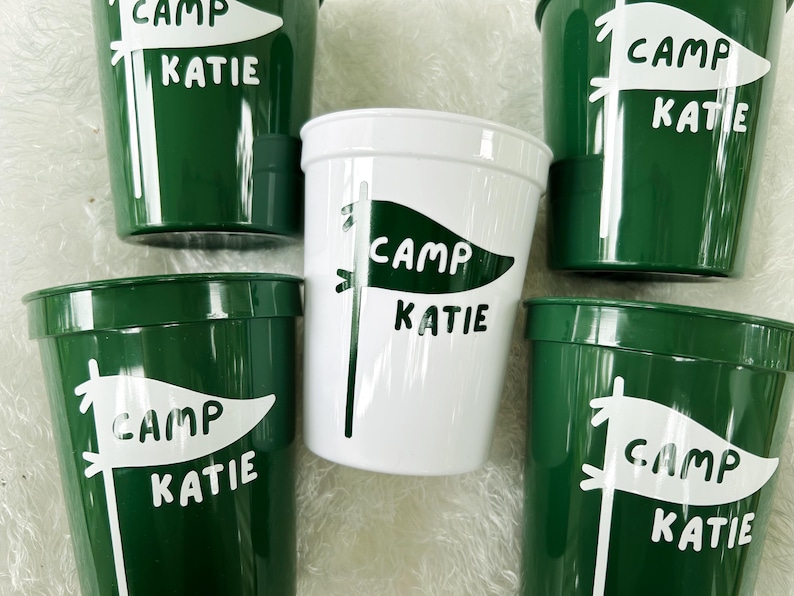 May include: Five green plastic cups with white lettering. Each cup has a white flag design with the text "Camp Katie" on it.