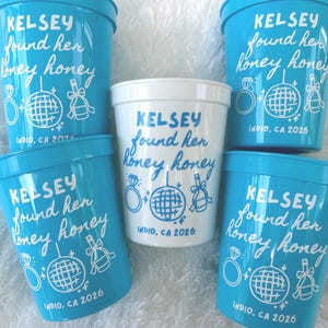 May include: Four bright blue plastic cups and one white plastic cup, each printed with the text KELSEY found her honey honey, a diamond ring icon, a disco ball, a champagne bottle, and the text INDIO, CA 2026 in a matching contrasting color.