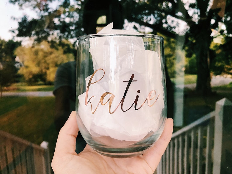 May include: A clear glass wine tumbler with the name "katie" written in gold lettering.