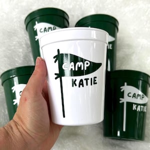 May include: Four green plastic cups with white lettering. One cup says "Camp Katie" with a white pennant design. Another cup says "Camp" with a white pennant design. Two cups say "Camp Katie" with a white pennant design.