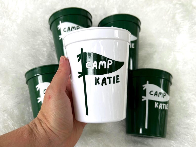 May include: Four green plastic cups with white lettering. One cup says "Camp Katie" with a white pennant design. Another cup says "Camp" with a white pennant design. Two cups say "Camp Katie" with a white pennant design.