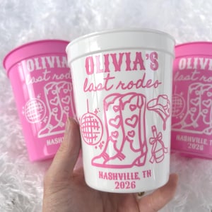 May include: Three plastic cups, two pink and one white, with a pink design. The cups feature the text "OLIVIA'S last rodeo" and illustrations of cowboy boots, a disco ball, and a cowboy hat. The text "NASHVILLE, TN 2026" is also present.