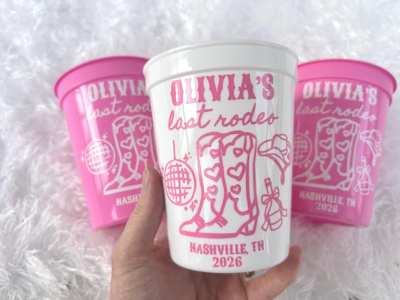 May include: Three plastic cups, two pink and one white, with a pink design. The cups feature the text "OLIVIA'S last rodeo" and illustrations of cowboy boots, a disco ball, and a cowboy hat. The text "NASHVILLE, TN 2026" is also present.
