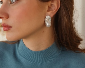 May include: A close-up of a person wearing large, irregular-shaped, pearl-colored stud earrings. The earrings are the focal point, with a textured, organic design. The person is wearing a teal-colored sweater.