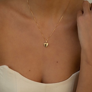 Puede incluir: Un collar con colgante de corazón dorado. El colgante en forma de corazón cuelga de una cadena fina. El collar se lleva sobre una parte superior de color claro.