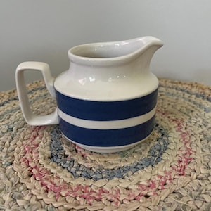 May include: A white ceramic creamer with two blue stripes around the middle. The creamer has a handle and a rounded shape. It is sitting on a woven rug.