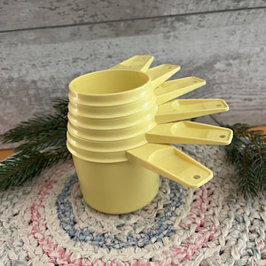 May include: A set of five nested pale yellow plastic measuring cups stacked vertically. Each cup features a flat handle with a small hole at the end, and the cups are graduated in size from the largest at the bottom to the smallest at the top.