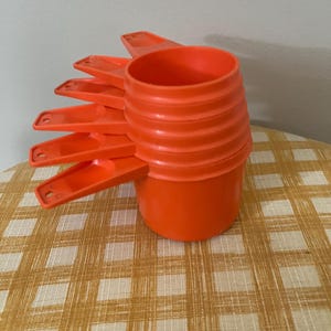 May include: A set of five bright orange plastic measuring cups stacked vertically. Each cup features a round bowl and a flat handle with a small circular hole at the end. The cups are nested together, creating a tiered appearance with uniform handles.