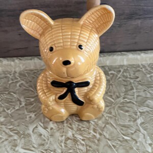 May include: A tan-colored ceramic bear-shaped container with a black bow tie. The bear has a textured surface, black eyes, and a small wooden handle on top. The container is sitting on a patterned surface.