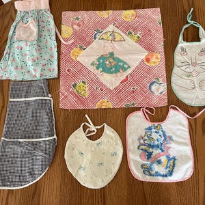 May include: A collection of vintage aprons and bibs in various patterns and colors. Includes a blue floral apron with a pink pocket, a black and white checkered apron, and several bibs with embroidered designs, including a cat and a floral pattern.
