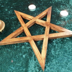 May include: A wooden pentagram, also known as a five-pointed star, with a dark brown stain finish. The pentagram is on a green velvet surface with two white candles.