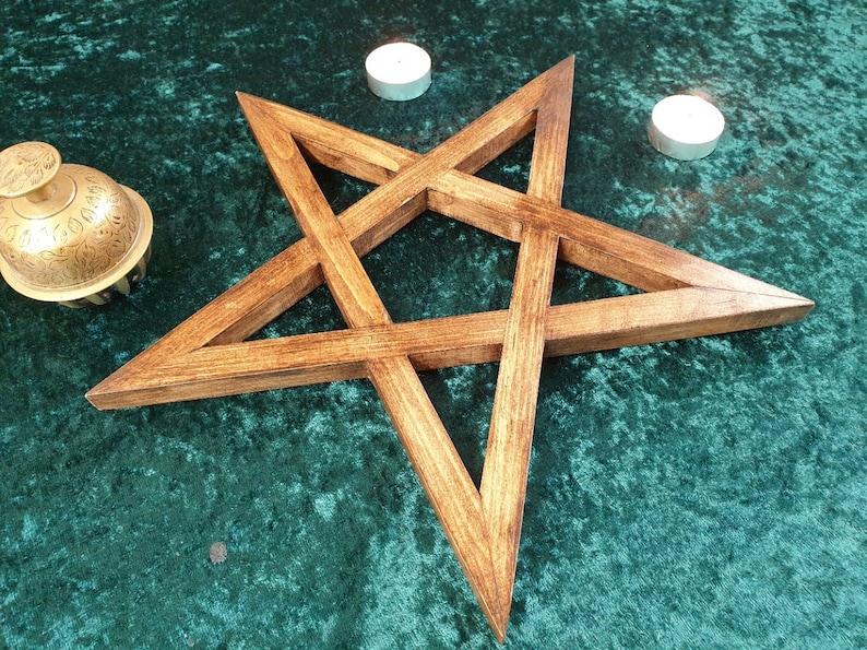 May include: A wooden pentagram, also known as a five-pointed star, with a dark brown stain finish. The pentagram is on a green velvet surface with two white candles.