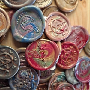 May include: A collection of colorful wax seals with various embossed designs, including pentagrams, crescent moons, and other mystical symbols. The seals feature a range of colors, such as red, blue, gold, and green, with a marbled effect. The seals are arranged on a wooden surface.