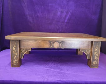May include: A dark brown wooden table with carved details. The table has a rectangular top and features a triple moon design and pentagrams. The legs are decorated with floral and leaf patterns. The table is set against a purple backdrop.