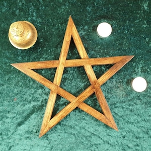 May include: A wooden pentagram, also known as a five-pointed star, on a green velvet background. Two white candles are placed on either side of the pentagram.
