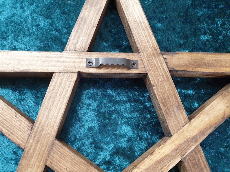May include: A wooden five-pointed star with a metal hanging loop on the back. The star is stained a dark brown color and has a rustic appearance.