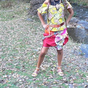 Puede incluir: Una mujer con rastas largas lleva un vestido estampado de colores con una cinta amarilla en la cintura. Está de pie en un entorno de bosque con un arroyo en el fondo.